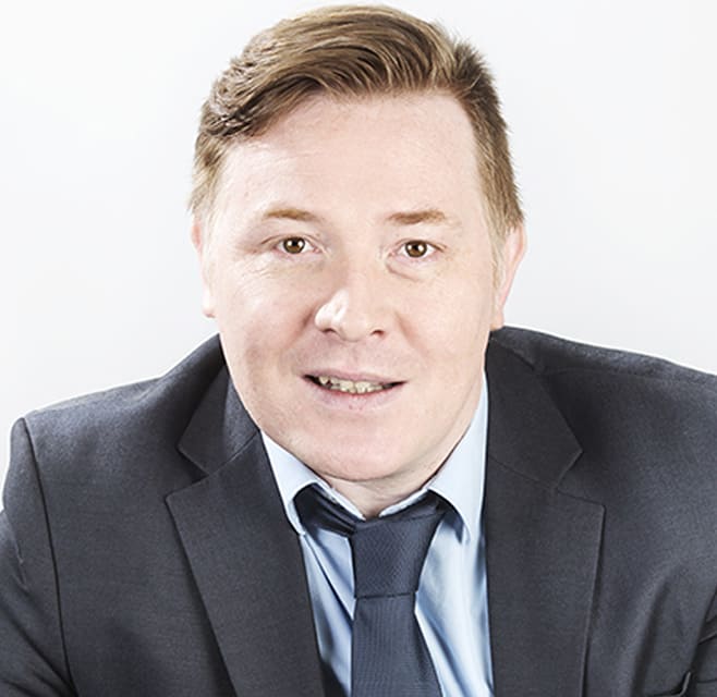 A man wearing a suit smiles into the camera. It's a studio setting.