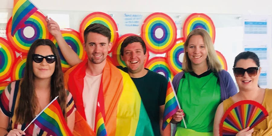 Cinq personnes sourient à la caméra, ils ont des drapeaux et des éventails pliants aux couleurs de l'arc-en-ciel.